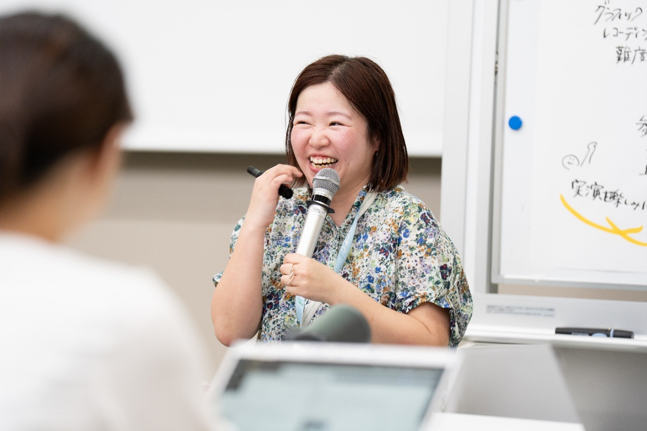 有廣さんが模造紙の前でマイクを持ち、参加者に向かって話している様子。笑顔。